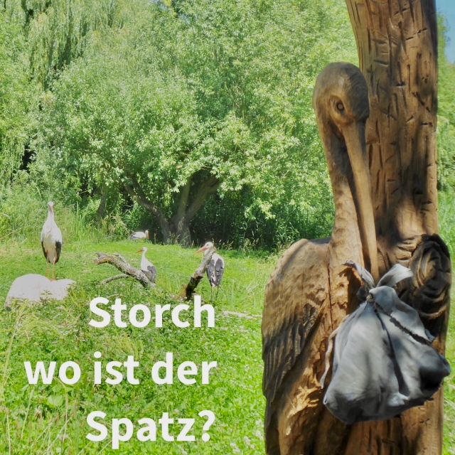 Holzplastik eines Storches mit Stoffbündel im Schnabel und Stöche auf einer Wiese mit Schriftzug Storch wo ist der Spatz